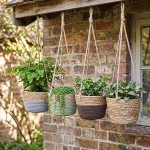 Può includere: Quattro vasi sospesi tramite spesse corde di iuta. Il primo è grigio e fibra naturale, il secondo è verde salvia e fibra naturale, il terzo è marrone scuro e fibra naturale, e il quarto è interamente realizzato in fibra di giunco intrecciata.
