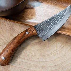 May include: A fixed-blade knife featuring a dark, hammered steel blade with a textured finish and a curved, polished wooden handle secured by two silver-toned metal rivets. The blade has a sharp, pointed tip and a curved cutting edge.