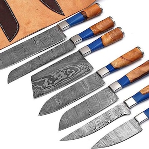 Può includere: Un set di sei coltelli da cucina con lame in acciaio Damasco dal motivo ondulato. Ogni coltello ha un manico in legno naturale e resina blu, con una ghiera color argento. I coltelli sono disposti in diagonale accanto a un rotolo in pelle.