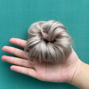May include: A round hair bun extension featuring synthetic hair in a light ash blonde shade with subtle platinum highlights. The hair is styled in a voluminous, textured circular shape with loose strands, designed to be worn as a hair accessory.