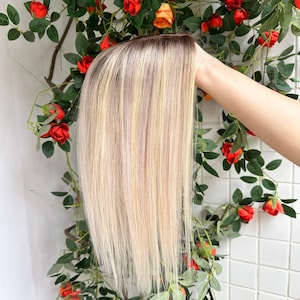 May include: A straight hair topper featuring a rooted design with dark brown roots transitioning into a blend of platinum blonde and light ash blonde highlights. The hair is sleek, smooth, and styled with a center part, showcasing a natural appearance.