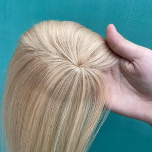 May include: A hair topper featuring straight, light blonde hair with a visible monofilament base at the crown. The hair is parted in the centre, showcasing a natural-looking scalp area where the individual strands are attached to the base.