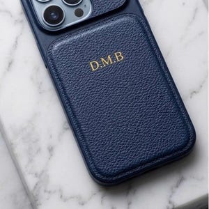 May include: A navy blue leather phone case with a textured finish. The case has a camera cutout and a flap with the gold initials "D.M.B." The case is on a white marble surface.