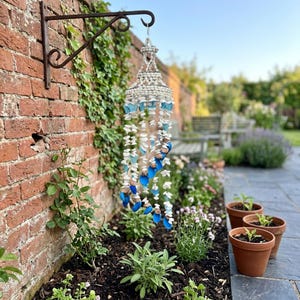 May include: A hanging wind chime constructed from white seashells and blue sea glass pieces arranged in multiple vertical strands suspended from a circular frame, attached to a dark metal wall bracket mounted on a red brick wall.