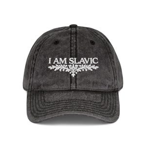 Könnte beinhalten: Eine anthrazitfarbene Baseballkappe mit verwaschenem, abgenutztem Finish. Auf der Vorderseite befindet sich der weiß gestickte Schriftzug I AM SLAVIC, der über einem dekorativen, symmetrischen, blumenartigen Ornament mittig platziert ist.