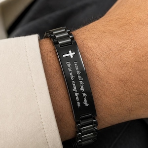 Op de afbeelding: Zwart roestvrijstalen armband met een kruissymbool en de inscriptie "Ik kan alles door Christus die mij sterkt." De armband wordt om een pols gedragen, met een wit overhemd en een grijs pak op de achtergrond.