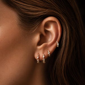 May include: Close-up of an ear adorned with multiple gold hoop earrings. Each earring is embellished with small, sparkling gemstones. The earrings are arranged in a cluster, creating a layered and elegant look. The skin tone is light.