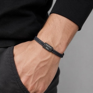 Peut inclure: Bracelet noir tressé avec un fermoir rectangulaire en métal gravé. Le fermoir porte les noms "Markus" et "Ella". Le bracelet est porté au poignet, mettant en valeur un design minimaliste.