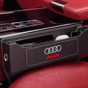 May include: A black leather car seat gap organizer featuring white contrast stitching and the silver Audi logo with red text. This rectangular storage accessory is positioned between the center console and a red leather car seat.