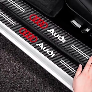 Peut inclure: Une protection de seuil de porte de voiture à texture fibre de carbone noire avec le logo Audi rouge et du texte blanc. La bande rectangulaire comprend deux rayures blanches horizontales à chaque extrémité, conçue pour le cadre de porte.