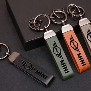 Puede incluir: Cuatro llaveros rectangulares con herrajes y anillas de metal oscuro. Cada uno tiene una correa en negro, verde oliva, tostado o gris, grabada con el logotipo y el texto MINI. Los llaveros están dispuestos con uno separado y tres sobre una caja.