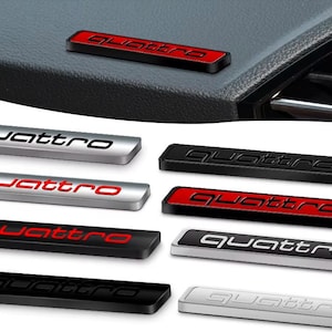 May include: A collection of rectangular automotive emblems featuring the word quattro in lowercase lettering. The badges are available in various finishes including brushed silver, matte black, and red, with some designs featuring contrasting colour inlays.