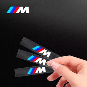 Op de afbeelding: Drie rechthoekige zwarte stickers met het M-logo en een ontwerp van schuine lichtblauwe, donkerblauwe en rode strepen. De stickers zijn in een waaier geplaatst, waarbij het iconische M-embleem duidelijk op elk afzonderlijk stuk zichtbaar is.