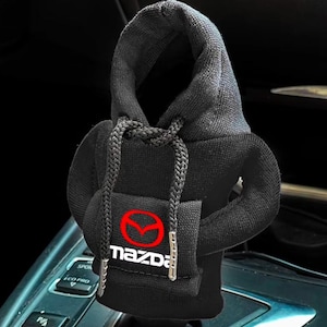 May include: A black fabric gear shifter cover designed like a miniature hoodie with a hood, long sleeves, and a front pocket. The front pocket features the red Mazda logo and the word "mazda" in white text. The hoodie includes black drawstrings.