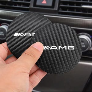 Könnte beinhalten: Zwei runde Auto-Getränkehalter-Einsätze mit schwarzem Carbonfaser-Strukturmuster. Jeder Einsatz zeigt das weiße AMG-Logo, das horizontal in der Mitte aufgedruckt ist. Die Untersetzer bestehen aus einem flexiblen Material für Fahrzeugkonsolen.