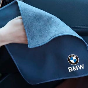 以下が含まれることがあります： 白、黒、青でBMWのロゴとテキストがプリントされた青いマイクロファイバー製のクリーニングクロス。布地は柔らかい質感の表面で縁が縫製されており、車両のダッシュボードの内装面を拭くために設計されています。