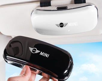 May include: A white and a black car visor sunglasses holder, each featuring a glossy finish and the MINI logo with the text MINI printed on the front. The white holder is attached to a car sun visor, while the black holder is held in the foreground.