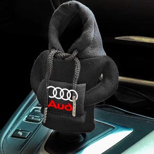 Könnte beinhalten: Eine schwarze Schalthebelabdeckung aus Stoff, gestaltet wie ein Miniatur-Kapuzenpullover mit Kapuze, langen Ärmeln und einer Vordertasche. Die Vordertasche zeigt das weiße Audi-Logo mit vier Ringen über dem roten Audi-Schriftzug auf dem Stoff.