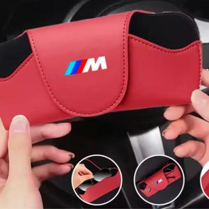May include: A red and black rectangular sunglasses case featuring the M logo in blue, purple, and red. The case has a magnetic flap closure. Three circular inset images show the case holding sunglasses, an open view, and the back clip attachment.