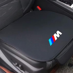 Op de afbeelding: Een zwart autostoelkussen met het M-logo en drie diagonale strepen in lichtblauw, donkerblauw en rood. Het kussen is gevormd om op de zitting te passen en is gemaakt van een glad, effen zwart materiaal met een strak afgewerkte rand.