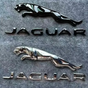 Puede incluir: Dos conjuntos de emblemas de la marca Jaguar, cada uno compuesto por una figura de jaguar saltando y la palabra JAGUAR en letras mayúsculas. Un conjunto tiene un acabado en negro brillante, mientras que el otro tiene una superficie metálica cromada.