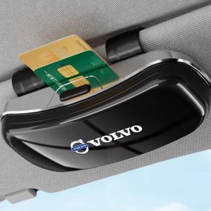 May include: A black and silver car visor clip featuring the Volvo logo and text. The accessory is attached to a gray fabric car sun visor and holds a gold and green credit card in its integrated slot, providing a secure storage solution.