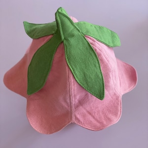 May include: A pink flower-shaped hat with green leaf accents. The hat is made of a soft, textured fabric and features a scalloped brim. The green leaves are attached to the top of the hat, adding a whimsical touch. This hat is perfect for a costume or everyday wear.