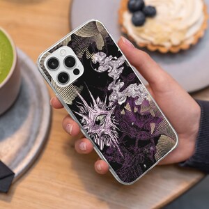 May include: A smartphone case featuring a black background with a detailed illustration of a spiky, eye-centered creature in shades of light purple and white, accented by swirling gold line patterns and wispy smoke-like shapes across the surface.