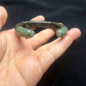 Könnte beinhalten: Ein dunkelgrünes, durchscheinendes Steinarmband mit C-förmigem Design und zwei einander zugewandten, geschnitzten Tierkopf-Endstücken. Die Oberfläche des Armbands weist ein texturiertes, segmentiertes Muster entlang des äußeren Bandes auf, das natürliche Farbvariationen und eine polierte Oberfläche zeigt.