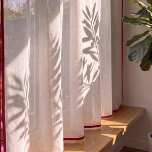 Può includere: Una tenda bianca semitrasparente con un bordo rosso vivace lungo i lati. Il tessuto presenta una trama visibile e la luce del sole proietta ombre distinte di rami di piante frondose sulla superficie della tenda appesa alla finestra.