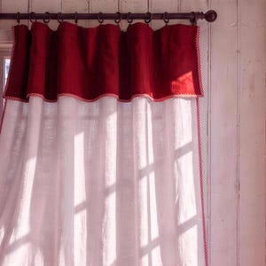 以下が含まれることがあります： 上部に深い赤色のバランス、縁に赤と白のストライプのパイピングが施された白いカーテンパネル。カーテンはリング付きの暗い色のロッドから吊り下げられており、日光が差し込み、窓枠の影がカーテンの表面に映し出されています。