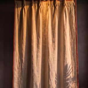 May include: A floor-length curtain made of light beige fabric with a textured weave. The curtain features vertical pleats at the top and is finished with a solid burnt orange border along the sides and bottom edges, casting a fern leaf shadow.