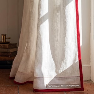 Op de afbeelding: Een wit linnen gordijnpaneel met een fel framboosrode rand langs de verticale zijde en de onderzoom. Een klein rechthoekig label onderaan bevat de tekst: Pure White Linen - Trimmed in Raspberry Red. De stof heeft een zichtbare weving.