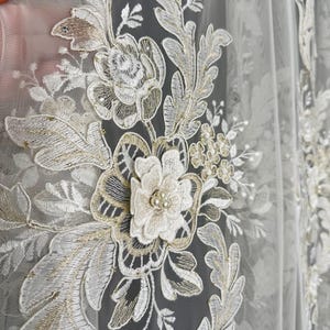 May include: Close-up of a sheer white fabric with detailed floral embroidery. The design features white and gold threadwork, including flowers, leaves, and pearl accents. The fabric is likely used for curtains or decorative purposes.