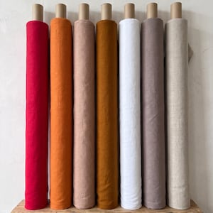 Op de afbeelding: Zeven verticale stofrollen op een rij, met een kleurenspectrum van felrood, oranje, lichtroze, mosterdgeel, wit, stoffig mauve en natuurlijk beige, allemaal gewikkeld rond kartonnen kokers en rechtopstaand geplaatst.
