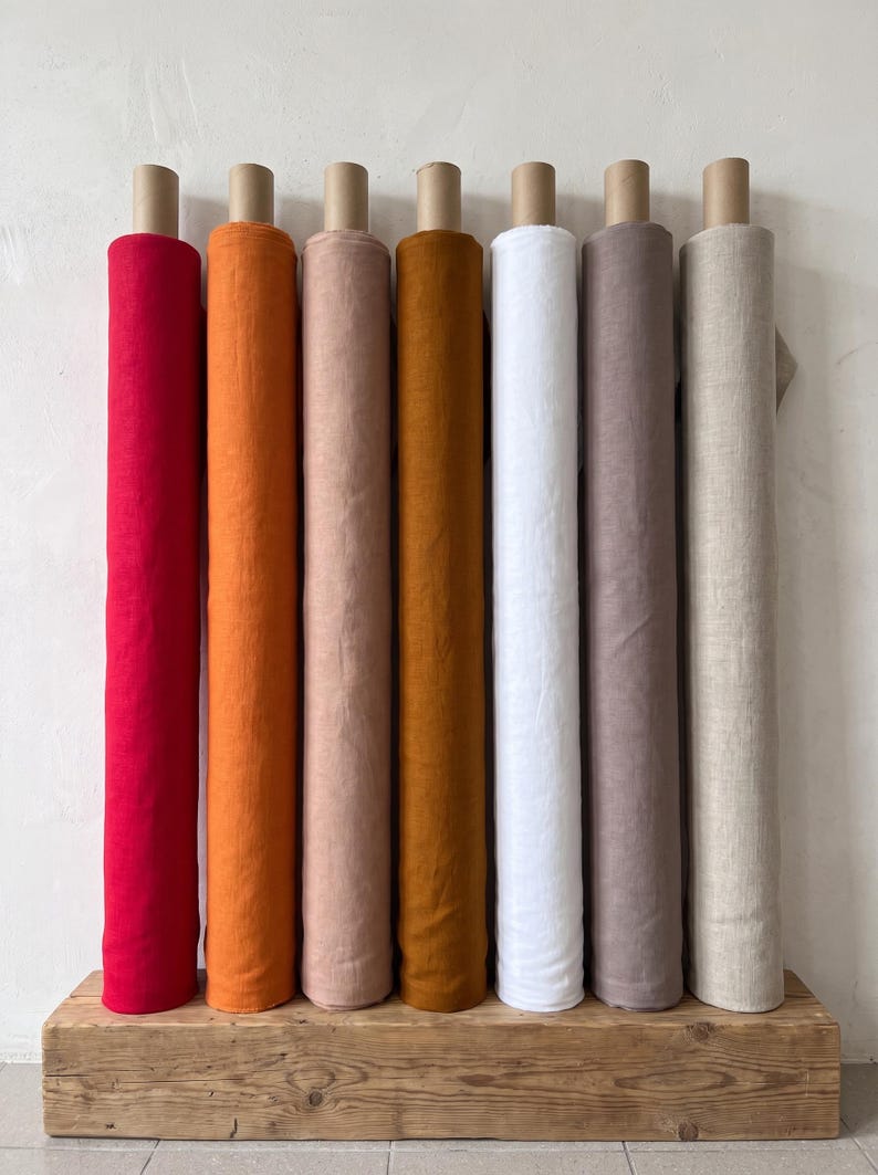 Op de afbeelding: Zeven verticale stofrollen op een rij, met een kleurenspectrum van felrood, oranje, lichtroze, mosterdgeel, wit, stoffig mauve en natuurlijk beige, allemaal gewikkeld rond kartonnen kokers en rechtopstaand geplaatst.
