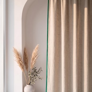 Op de afbeelding: Een beige gordijn met een felgroene randafwerking dat verticaal hangt. Naast het gordijn staat een lichtgekleurde keramische vaas met drie hoge, vederachtige pampasgrasstengels en een kleine tak met groene bladeren, geplaatst in een boogvormige nis.