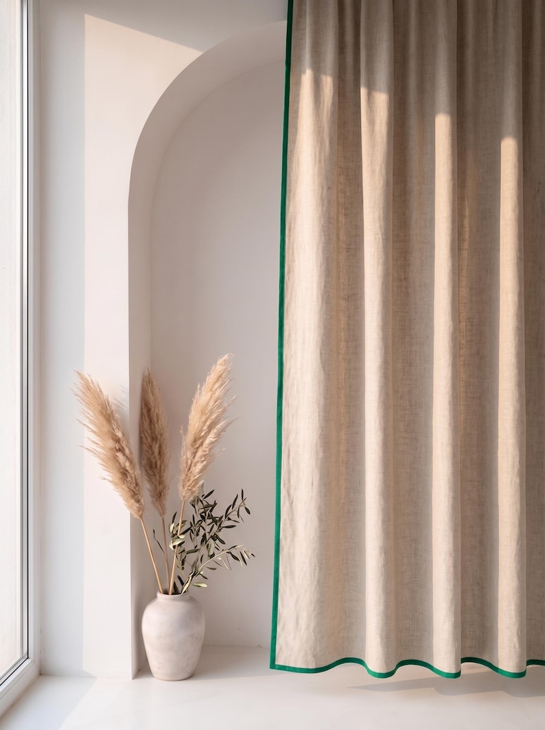 Op de afbeelding: Een beige gordijn met een felgroene randafwerking dat verticaal hangt. Naast het gordijn staat een lichtgekleurde keramische vaas met drie hoge, vederachtige pampasgrasstengels en een kleine tak met groene bladeren, geplaatst in een boogvormige nis.