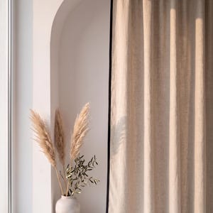 Op de afbeelding: Een kamerhoog beige gordijn met een effen zwarte randafwerking dat verticaal hangt. Naast het gordijn staat een lichtgekleurde keramische vaas met verschillende hoge, vederachtige pluimen van pampasgras en een tak met groene bladeren.