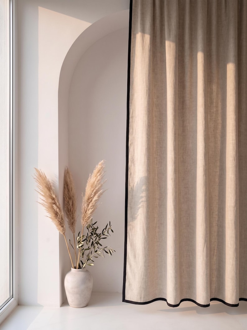 Op de afbeelding: Een kamerhoog beige gordijn met een effen zwarte randafwerking dat verticaal hangt. Naast het gordijn staat een lichtgekleurde keramische vaas met verschillende hoge, vederachtige pluimen van pampasgras en een tak met groene bladeren.