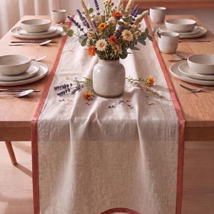 Op de afbeelding: Een beige linnen tafelloper met een terracotta-kleurige rand over een houten eettafel. In het midden staat een keramieken vaas met een boeket van gedroogde lavendel, oranje bloemen en grassen, omringd door verschillende tafeldekkingen.