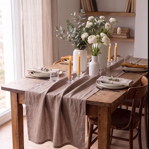 Pode incluir: Uma mesa de jantar de madeira rústica posta para uma refeição com um caminho de mesa de linho castanho claro. A mesa apresenta vasos de cerâmica branca cheios de flores brancas e eucalipto, velas amarelas em suportes de vidro e pratos.