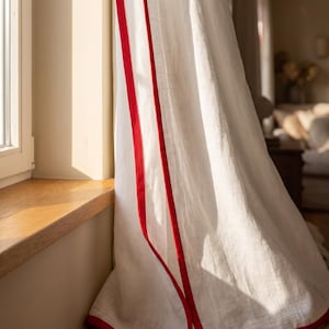 Op de afbeelding: Een wit gordijn met een felrode stoffen rand langs de verticale zijde en de onderzoom. De stof heeft een zichtbare geweven textuur en hangt in zachte plooien, waarbij het natuurlijke licht bij een houten vensterbank wordt opgevangen.