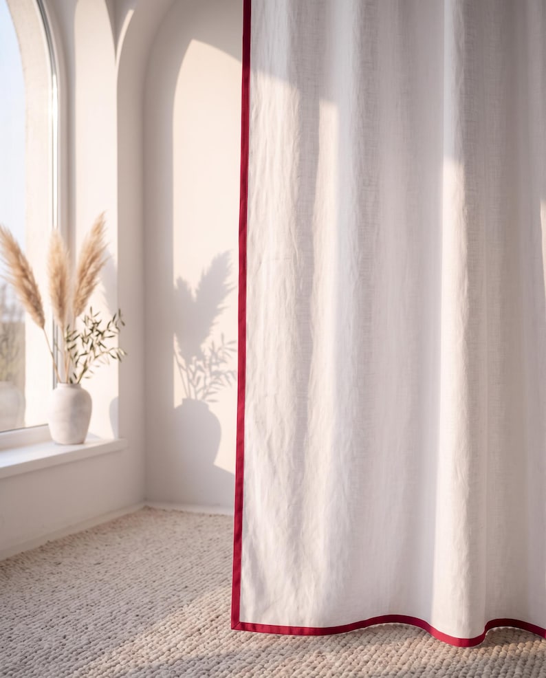 Op de afbeelding: Een kamerhoog wit gordijn met een effen dieprode rand langs de verticale en onderste zijden. De stof heeft een zichtbare geweven textuur en hangt in zachte, verticale plooien, wat zorgt voor een strakke en gestructureerde uitstraling.