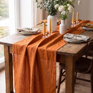 May include: A long, burnt orange linen table runner draped over a rustic wooden dining table. The table is set with white ceramic plates, beige linen napkins, white ribbed vases containing white flowers, and yellow beeswax taper candles.