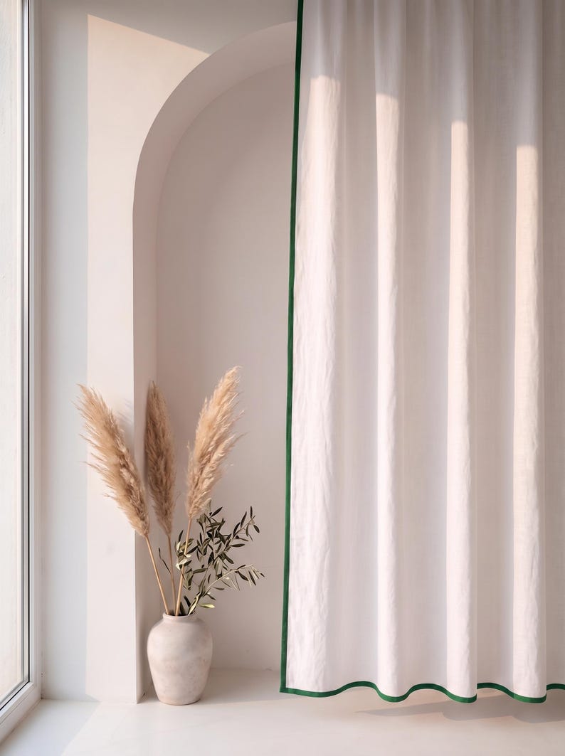 Op de afbeelding: Een kamerhoog wit gordijn met een donkergroene rand langs de zijkanten. Naast het gordijn staat een lichtgekleurde keramische vaas met drie hoge, gevederde pampasgrasstengels en een kleine tak met groene olijfbladeren.