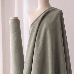 Könnte beinhalten: Eine Stoffrolle in Salbeigrün, die über eine Schneiderpuppe drapiert ist. Das Textil weist eine sichtbare Webstruktur mit matter Oberfläche auf und zeigt eine gleichmäßige Farbe. Der Stoff ist links um einen zylindrischen Kern gewickelt.