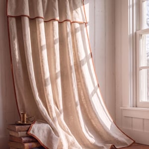 Può includere: Una tenda color beige a tutta altezza con mantovana superiore e un bordo a contrasto color ruggine lungo i lati. La tenda è appesa a un bastone in metallo scuro con anelli e cade formando delle pieghe verticali lungo la finestra.