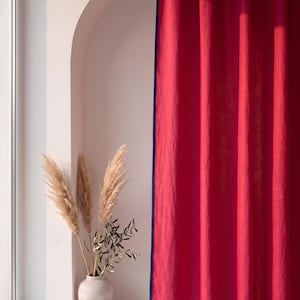 Op de afbeelding: Een levendig magentakleurig gordijn met een donkerblauwe rand langs de verticale en onderste randen. De stof heeft een zichtbare textuur en hangt in verticale plooien. Naast het gordijn staat een lichtgekleurde vaas met gedroogd pampasgras.