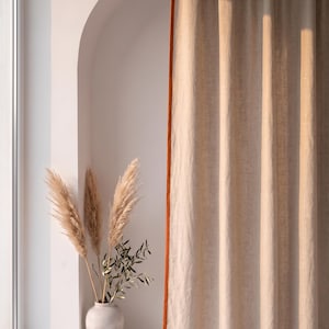 Op de afbeelding: Een kamerhoog beige gordijn met een feloranje randafwerking dat verticaal hangt. Naast het gordijn staat een lichtgekleurde keramische vaas met een verzameling hoge, vederachtige pampasgraspluimen en een takje met groene olijfbladeren.